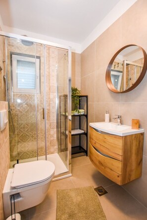 Ein modernes Badezimmer mit einer Duschkabine aus Glas, einer weißen Toilette, einem hölzernen Waschtisch mit Spiegel und einer kleinen Pflanze auf dem Regal.
