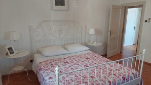Ein Schlafzimmer mit einem weißen Bett, einer blumigen Tagesdecke, zwei Nachttischen mit Lampen, einem Bilderrahmen und einer Tür zu einem anderen Raum.