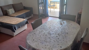 Un coin repas avec une table ronde recouverte d’une nappe à motifs floraux, entourée de chaises. On aperçoit, au fond, un canapé avec des coussins et un oreiller.