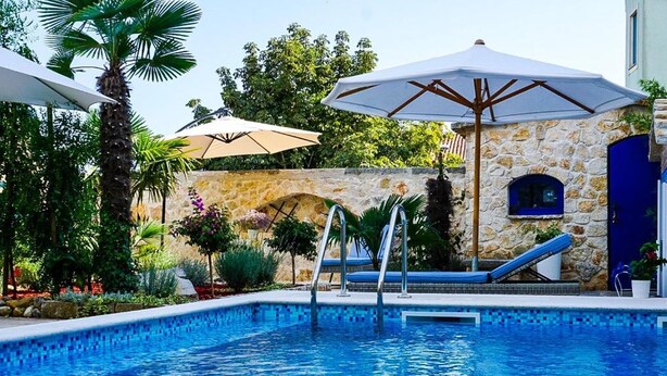 Ein Schwimmbecken mit Steinmauer und Palmen. Liegestühle und Sonnenschirme sind vorhanden.