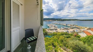 A balcony with a chair, a glass table, and a view of a marina with boats and a coastal town. - Apartments Sanja (Rab)