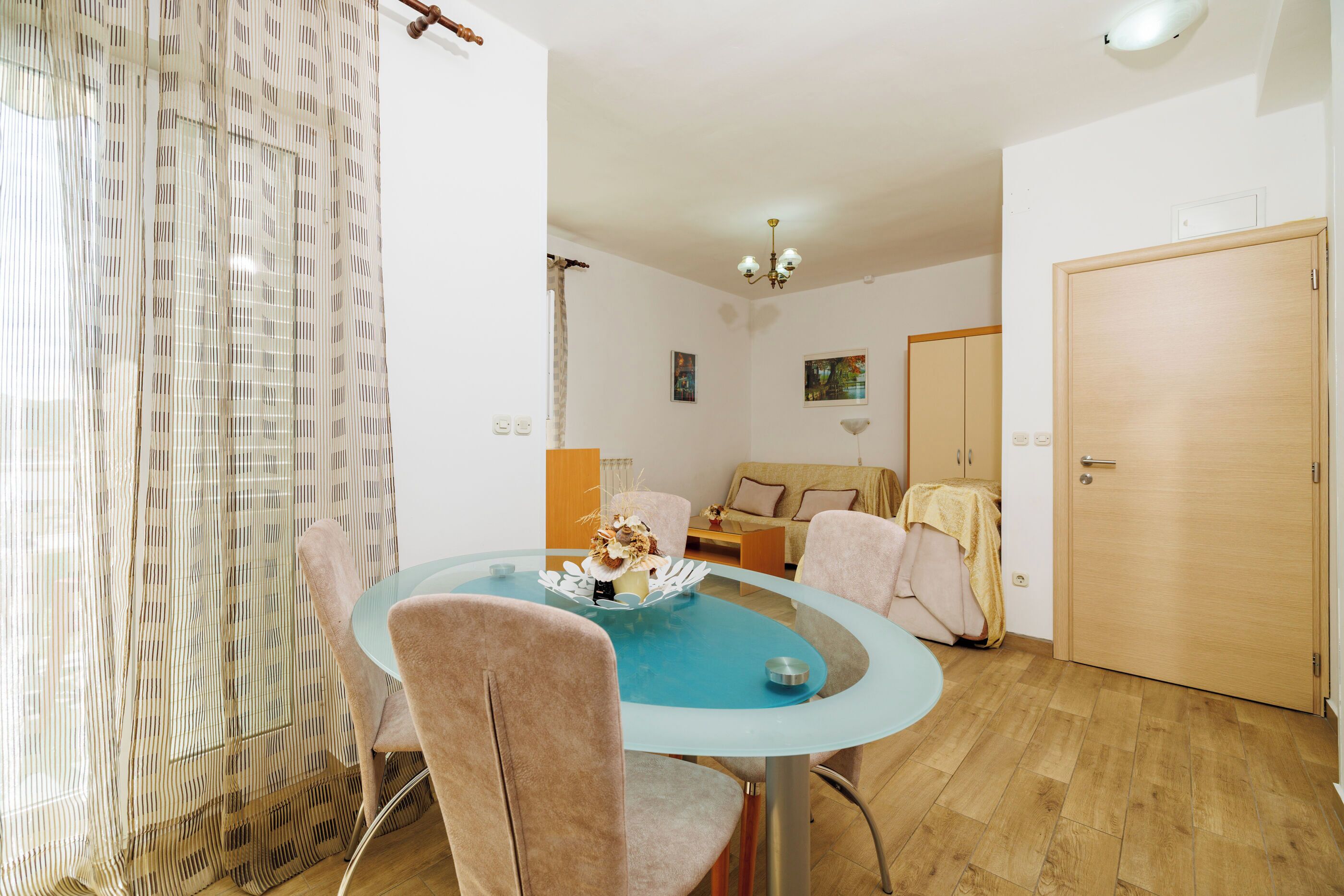 A room with a round glass table, two chairs, a sofa, a wooden door, and a window with sheer curtains.