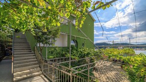 A green building with a staircase leading to a balcony, surrounded by greenery and overlooking a body of water. - Apartments Sanja (Rab)
