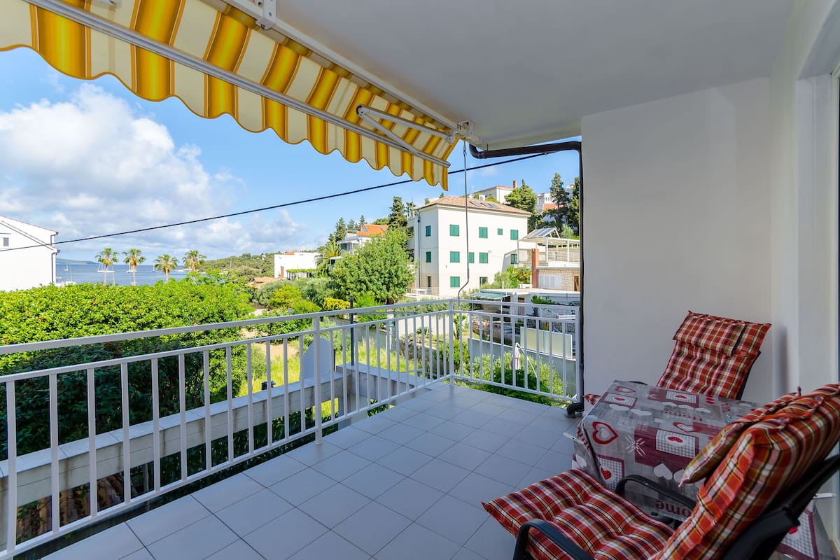 A balcony with a white railing, a yellow awning, and a view of the sea and buildings in the background.