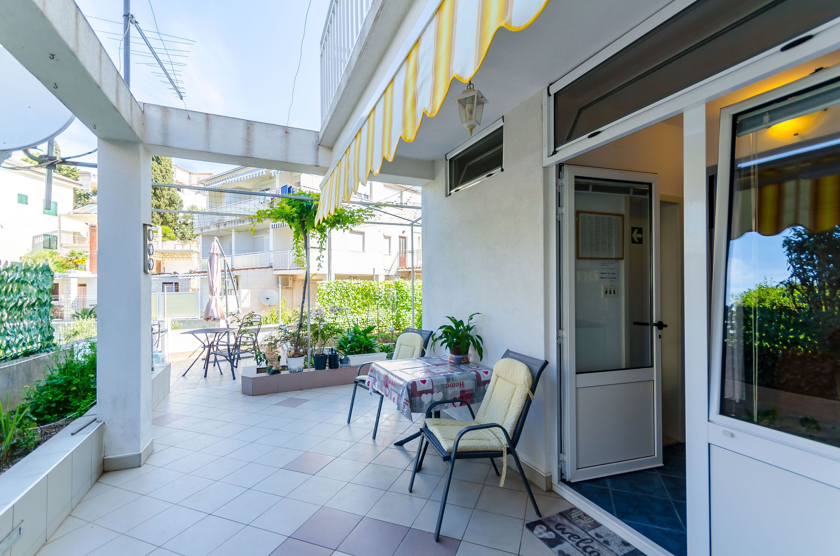 A covered outdoor patio with a table and chairs, potted plants, and a view of neighboring buildings.