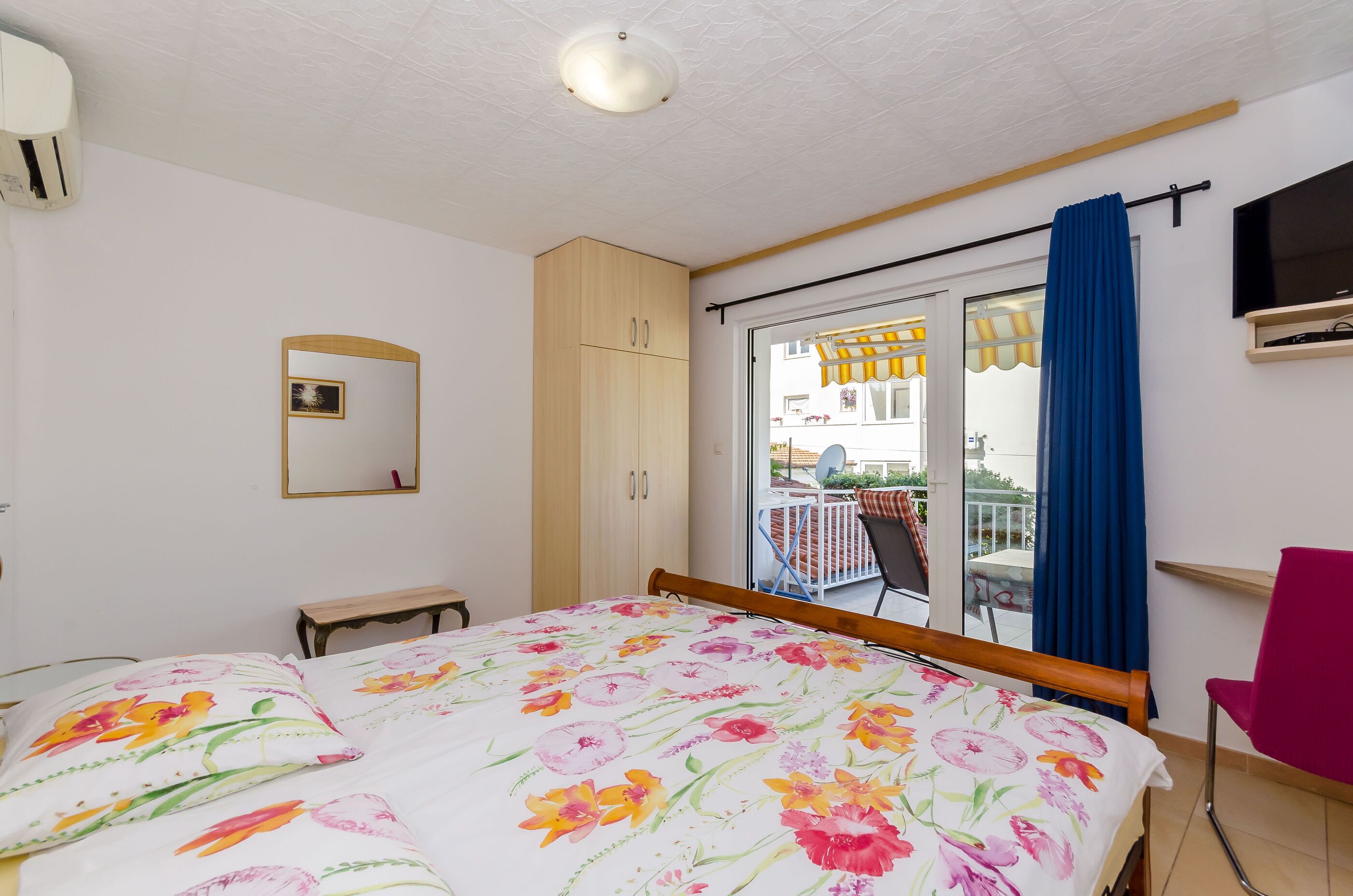 A bedroom with a bed, a wooden wardrobe, a small table, a mirror, a TV, and a balcony with a blue curtain.
