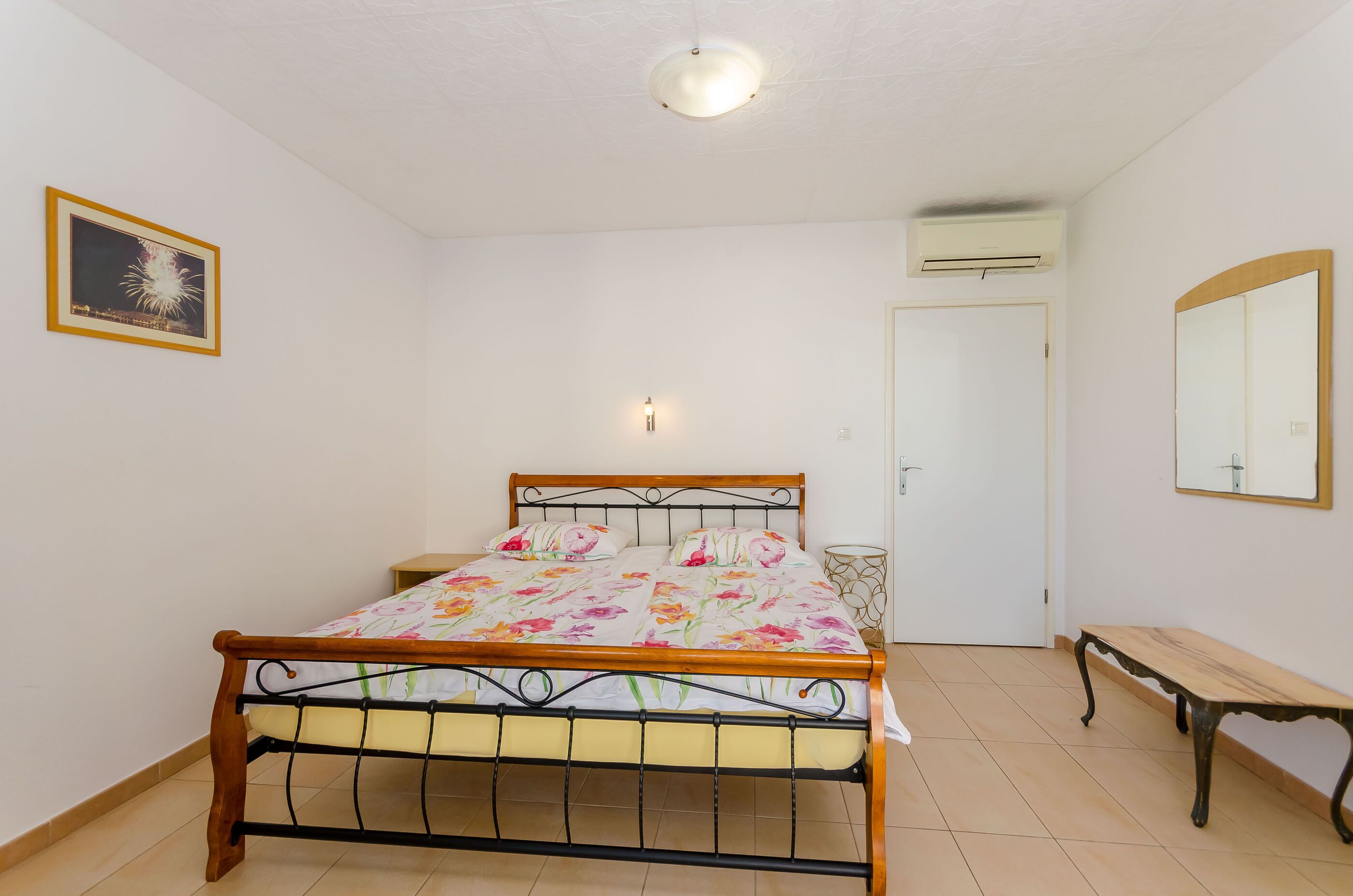 A bedroom with a wooden bed, a bedside table, a small round table, a bench, a framed picture on the wall, and an air conditioning unit.
