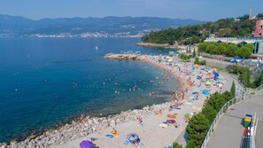 Ein Küstenstrand mit klarem, blauem Wasser, felsiger Küstenlinie und einer Promenade mit Sonnenschirmen und Badegästen.