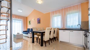 A modern living room with a sofa, dining table, and chairs, a kitchen with white cabinets, and a staircase with a wooden railing. - Apartments Kostrena (Kostrena)