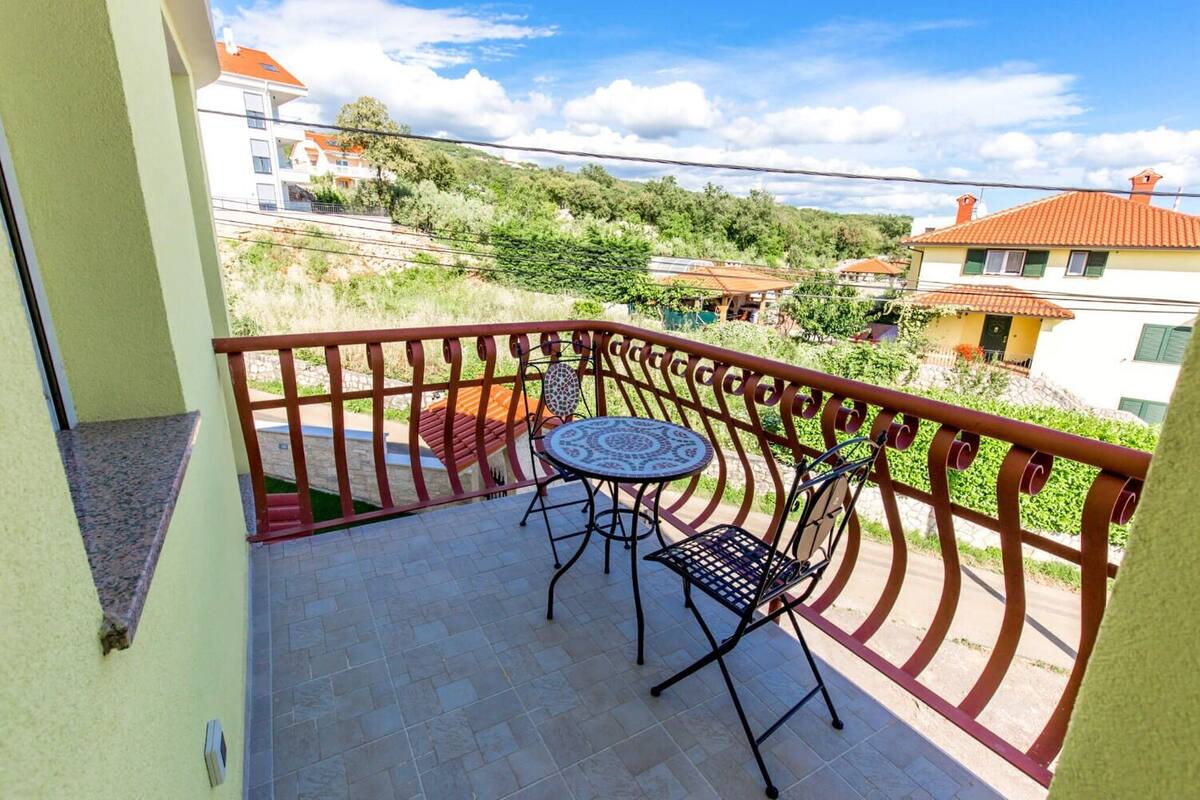 A balcony with a table and chairs, a red railing, and a view of houses and greenery.