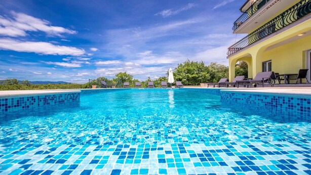 A clear blue pool with tiled edges, surrounded by lounge chairs and a yellow building with a balcony. - Apartments Kostrena (Kostrena)