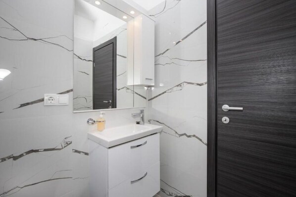 A modern bathroom with a white vanity, a mirror, and a dark wooden door. - Apartments Jakov (Postira)