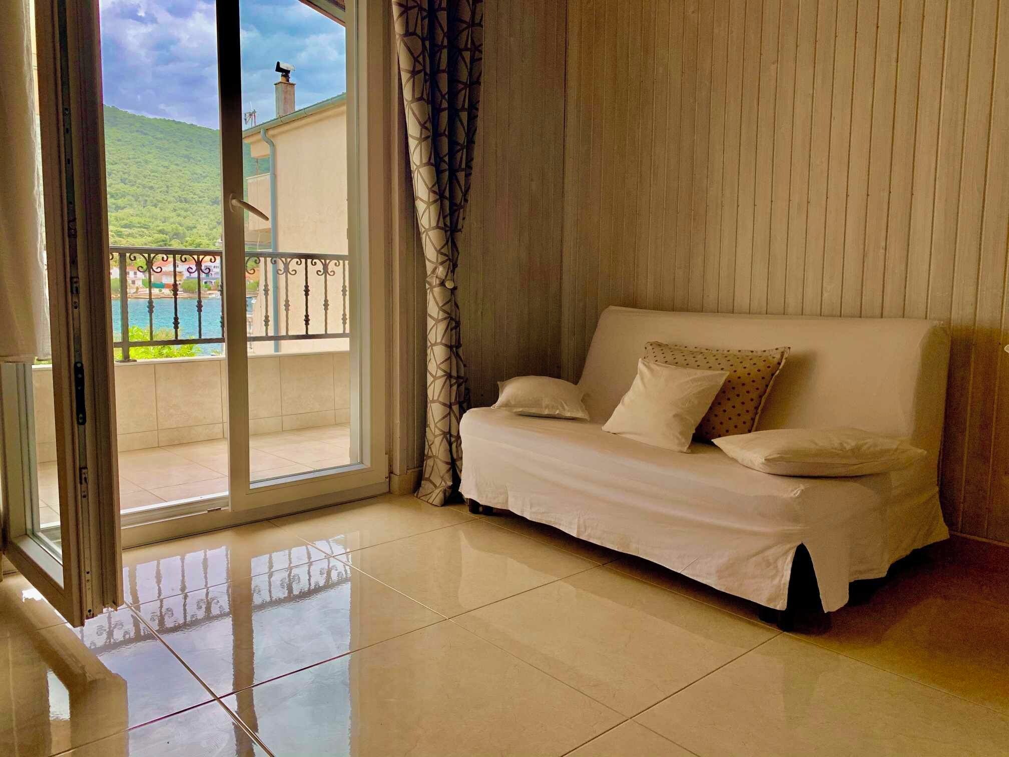 Una habitación con un sofá blanco, un ventanal corredero que da a un balcón y vistas a un cuerpo de agua y follaje.