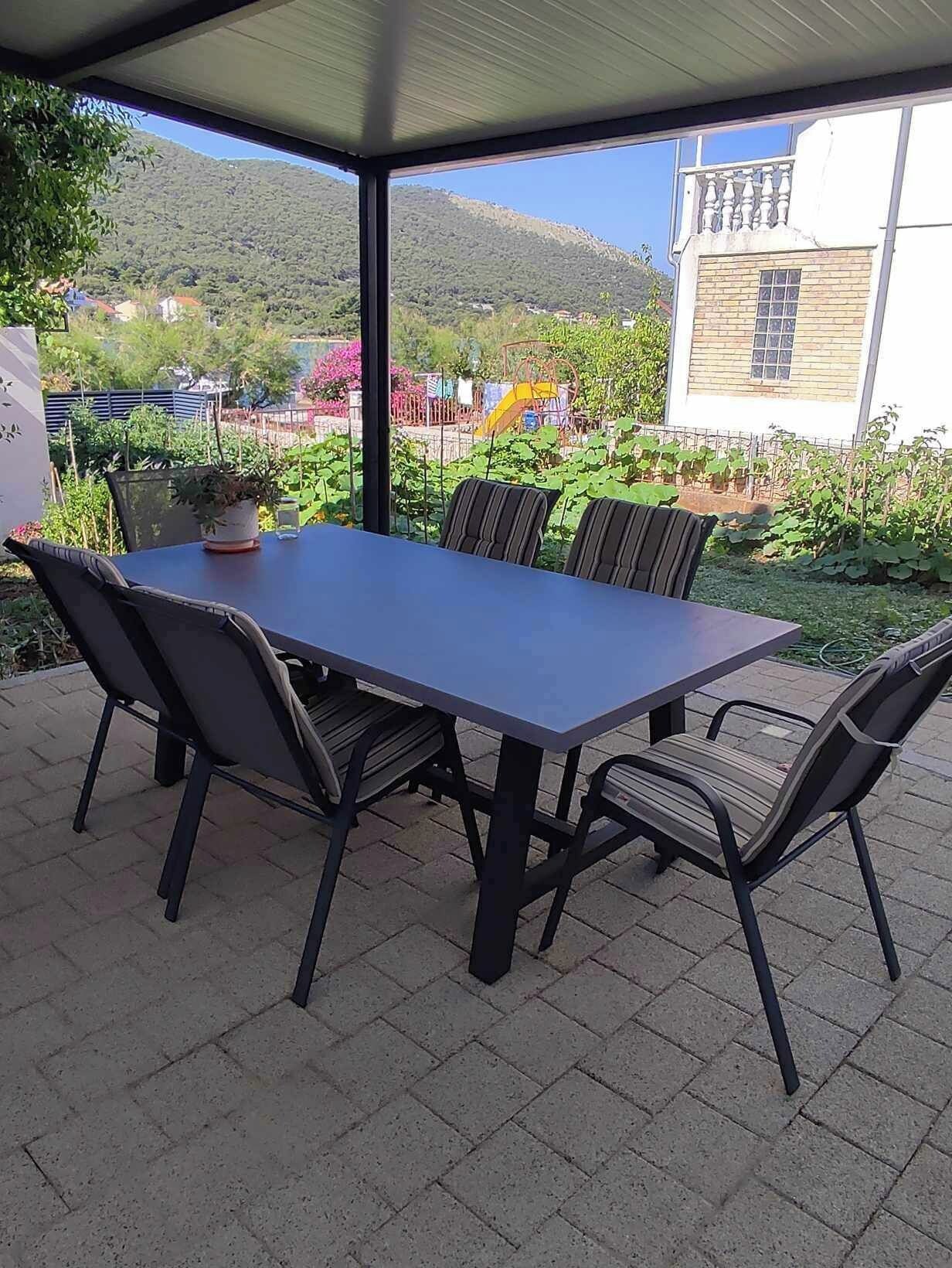 A covered patio with a rectangular table and six chairs, surrounded by a garden with various plants and a playground in the background.
