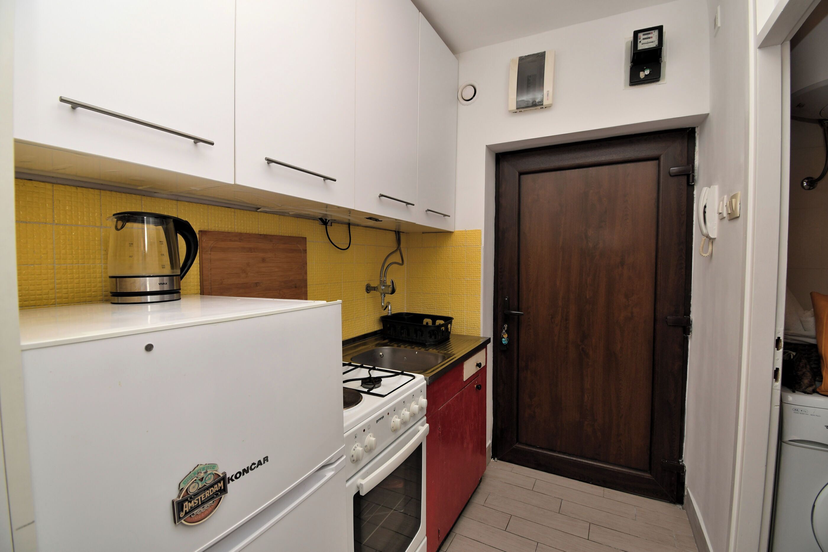 Une cuisine de taille réduite avec des meubles blancs, une cuisinière à gaz, une machine à café et un évier rouge.