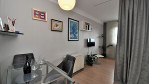 A modern living room with a glass dining table, a black sofa, a beige cabinet, and a flat-screen TV mounted on the wall. - Apartment City (Rijeka)
