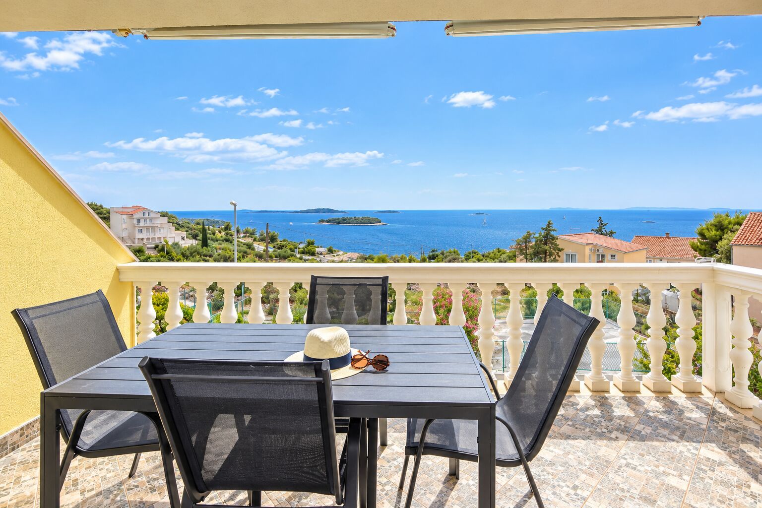 A balcony with a table and chairs, overlooking a coastal view with houses and the sea.