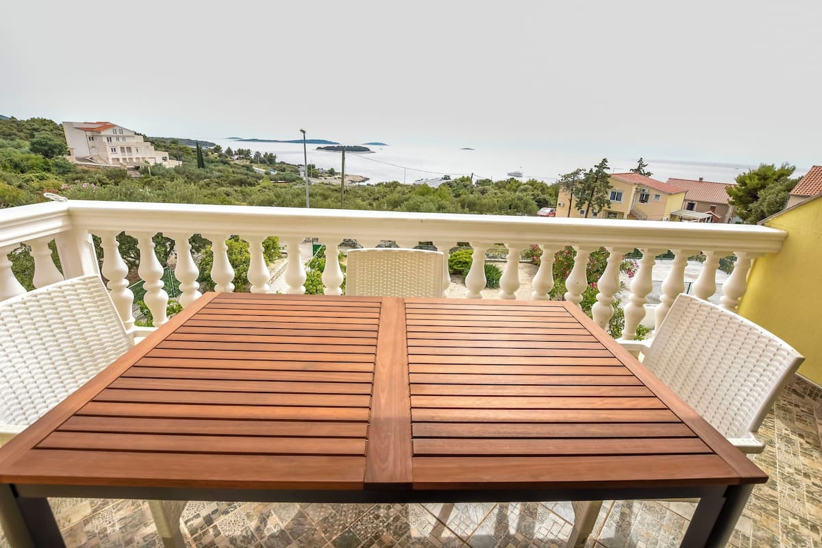 A wooden table and white chairs on a balcony with a view of the sea and buildings.