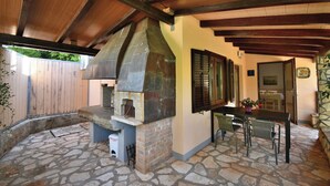 Un patio al aire libre con suelo de piedra, horno de ladrillo, vigas de madera, una mesa con sillas y una planta en maceta.