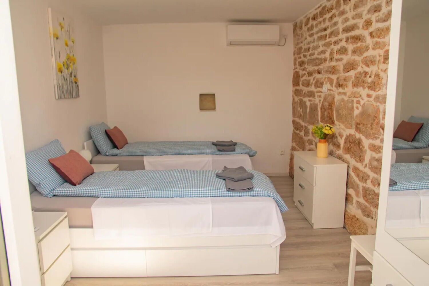 A room with two beds, each with a checkered bedspread and pillows. There is a white dresser and a small table with a vase of flowers. The room has a stone wall and an air conditioning unit on the wall.
