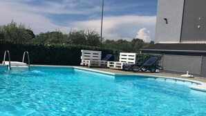 A swimming pool with clear blue water, surrounded by white lounge chairs and a tall building in the background. - Apartment Mirella (Umag)