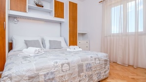 Ein ordentlich bezogenes Bett mit gemustertem Überwurf, ein Holzschrank, ein kleiner Nachttisch und ein Fenster mit durchscheinenden Vorhängen.