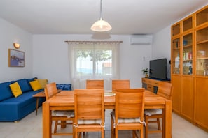 Un coin repas avec une table et des chaises en bois, un canapé bleu avec des coussins jaunes, un téléviseur posé sur un meuble et un buffet en bois avec des portes en verre.