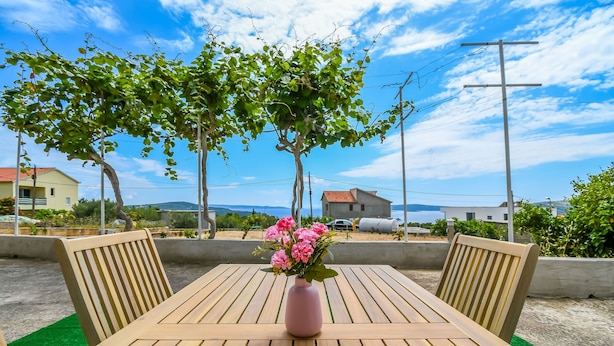 Ein Holztisch mit einer rosa Blumenvase, zwei Holzstühle und im Hintergrund eine Aussicht auf Häuser und Bäume.