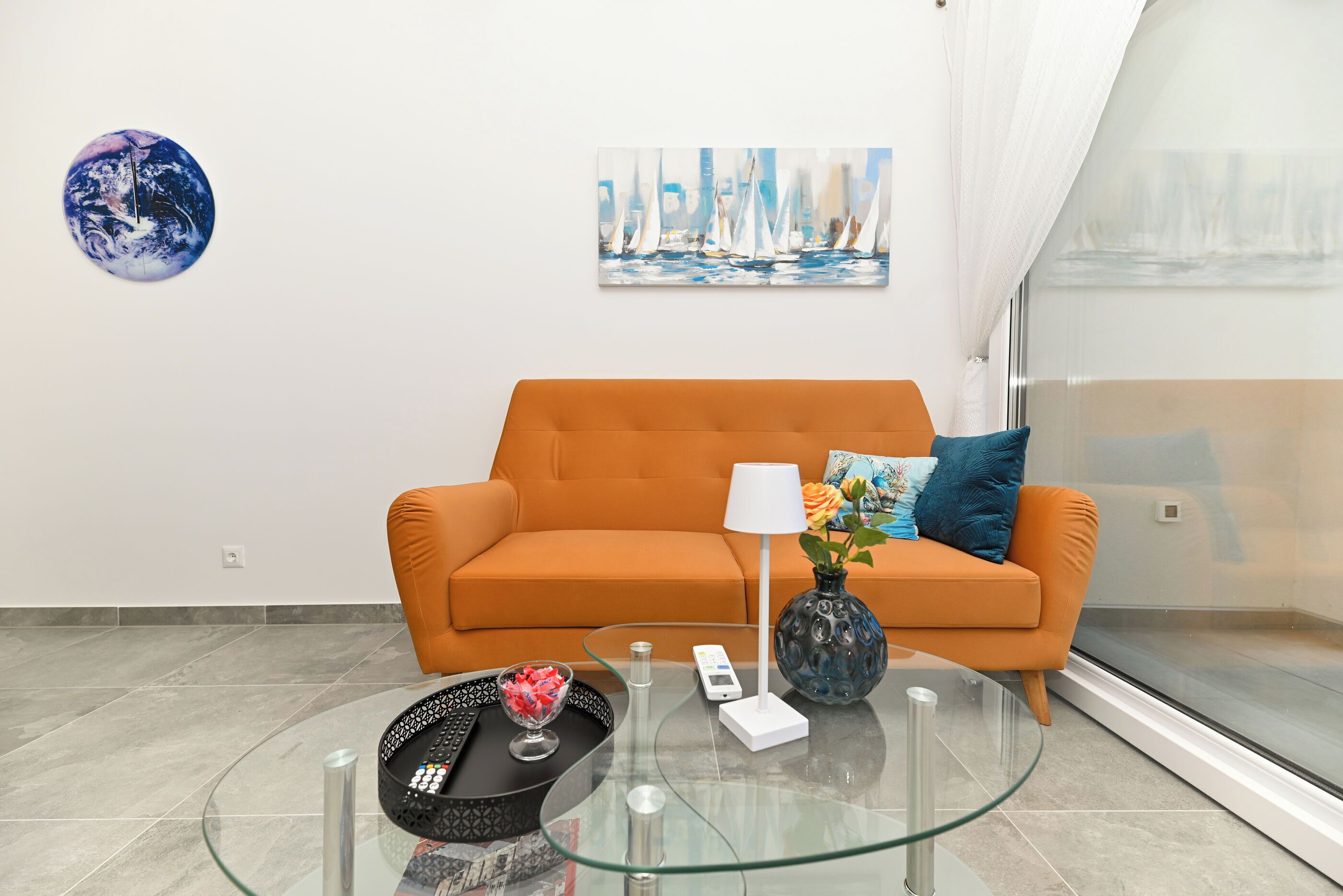 En moderne stue med et glasbord, en orange sofa og et maleri af sejlbåde.