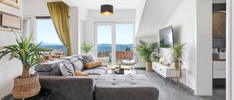 A modern living room with a grey sectional sofa, a round coffee table, and a large window with a view of the sea.