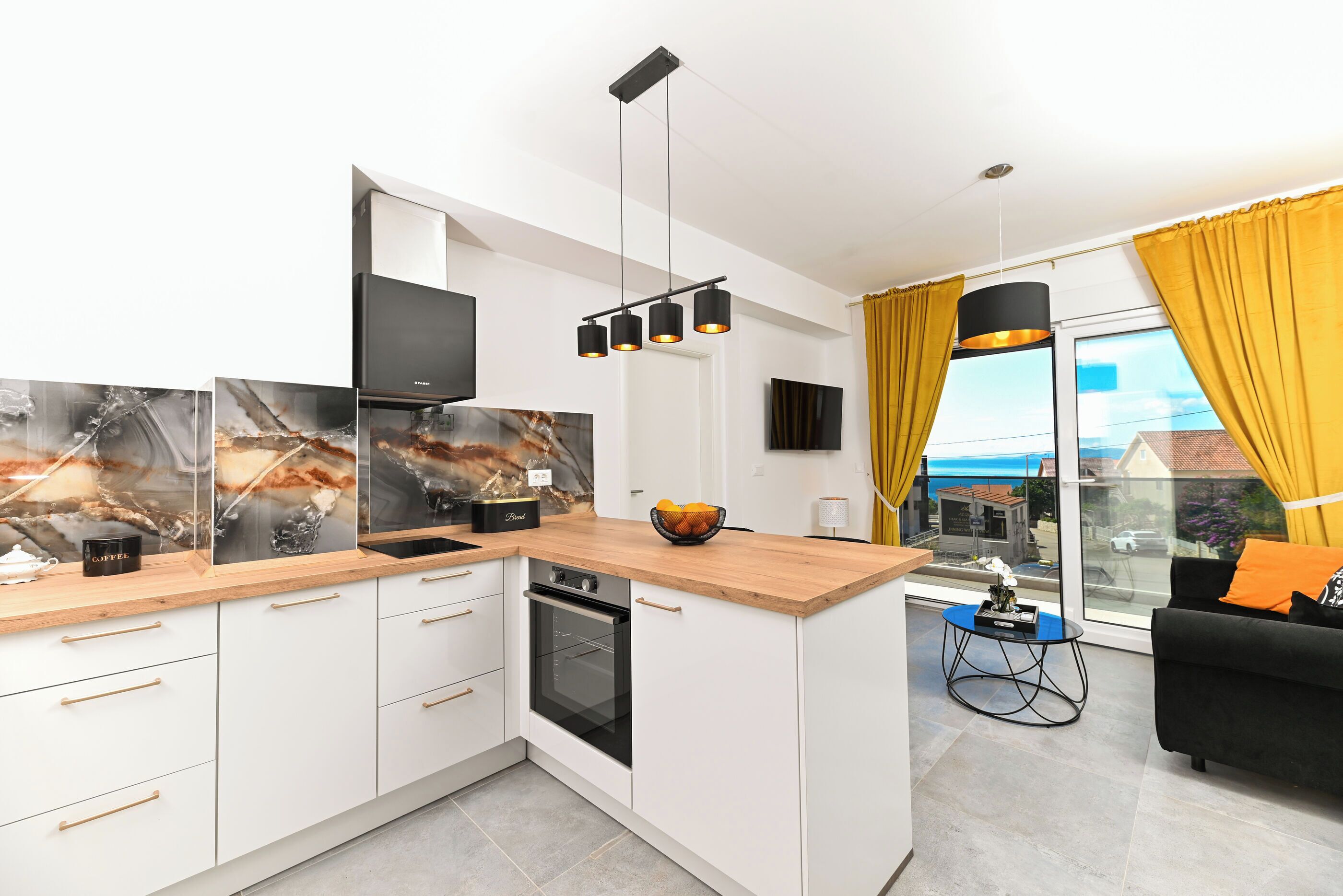 A modern kitchen with white cabinets, a wooden countertop, and a built-in oven. There is a black coffee machine on the counter, and a bowl of fruit. The kitchen has a large window with yellow curtains and a view of the outside.