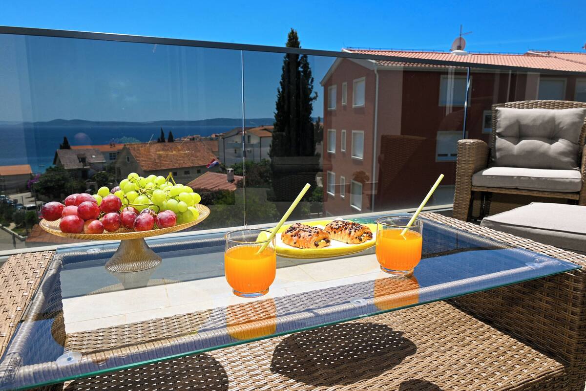 A balcony with a glass table displaying a fruit platter, two glasses of orange juice, and a wicker chair.