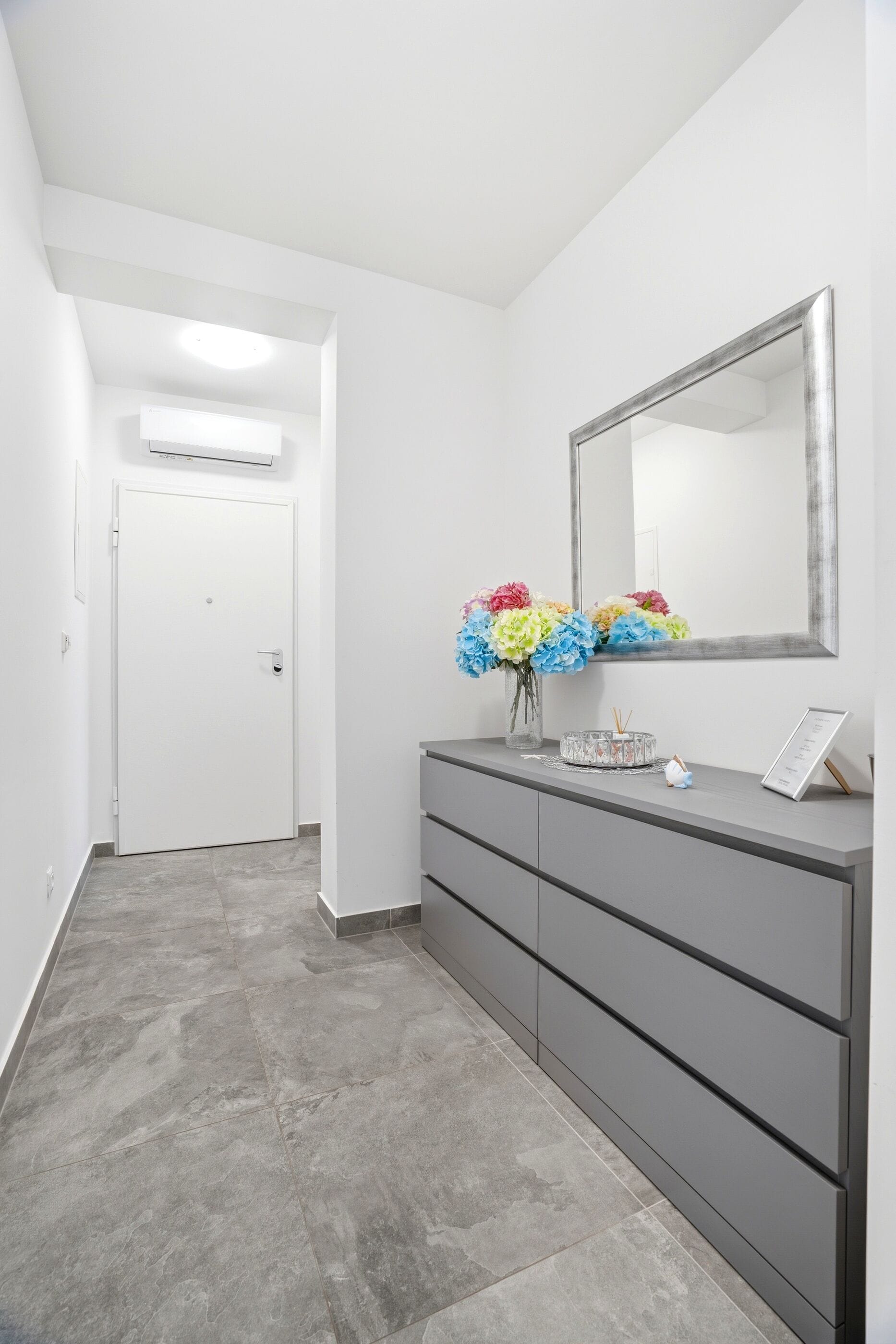 Un couloir avec une commode grise, un miroir et un vase de fleurs.