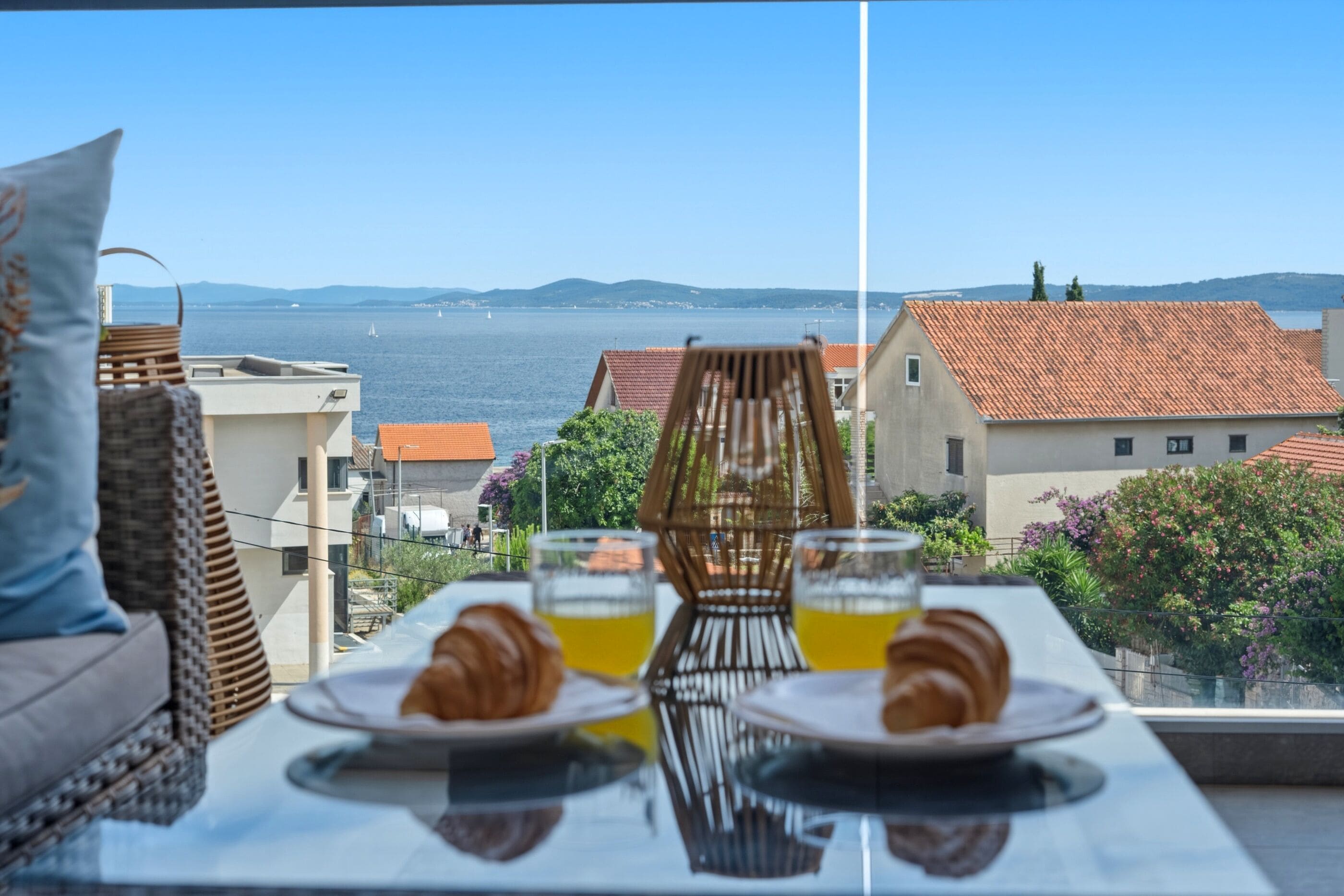 A table with plates, glasses, and a croissant, overlooking a coastal view with buildings and a clear sky.