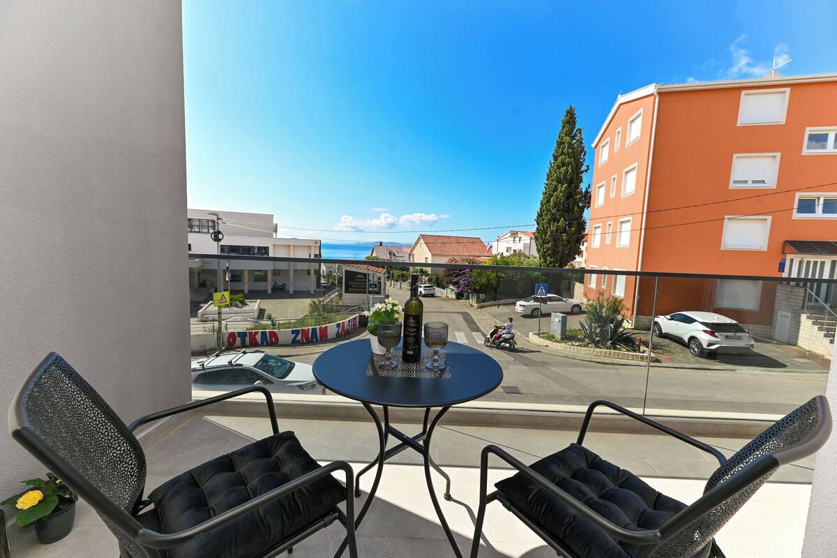 A balcony with two chairs and a small table, overlooking a street with buildings and parked cars.