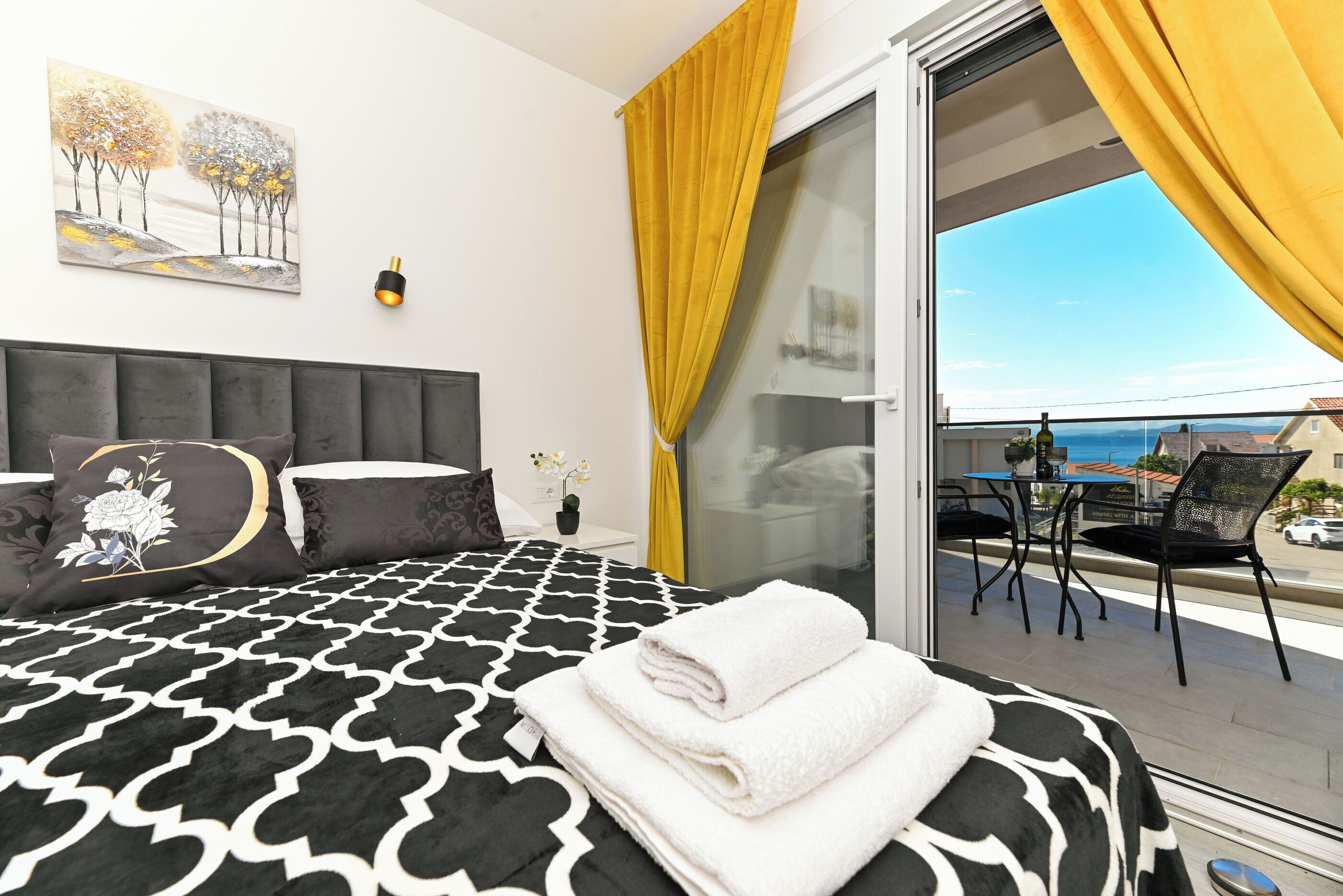 A bedroom with a black and white patterned bedspread, a grey upholstered headboard, a painting of trees, a small bedside table with a vase of flowers, and a balcony with yellow curtains and a view of the sea.