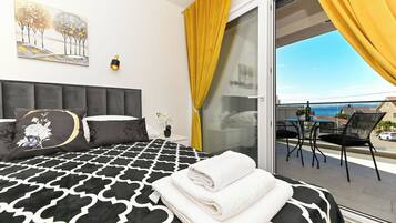A bedroom with a black and white patterned bedspread, a grey upholstered headboard, a painting of trees, a small bedside table with a vase of flowers, and a balcony with yellow curtains and a view of the sea.