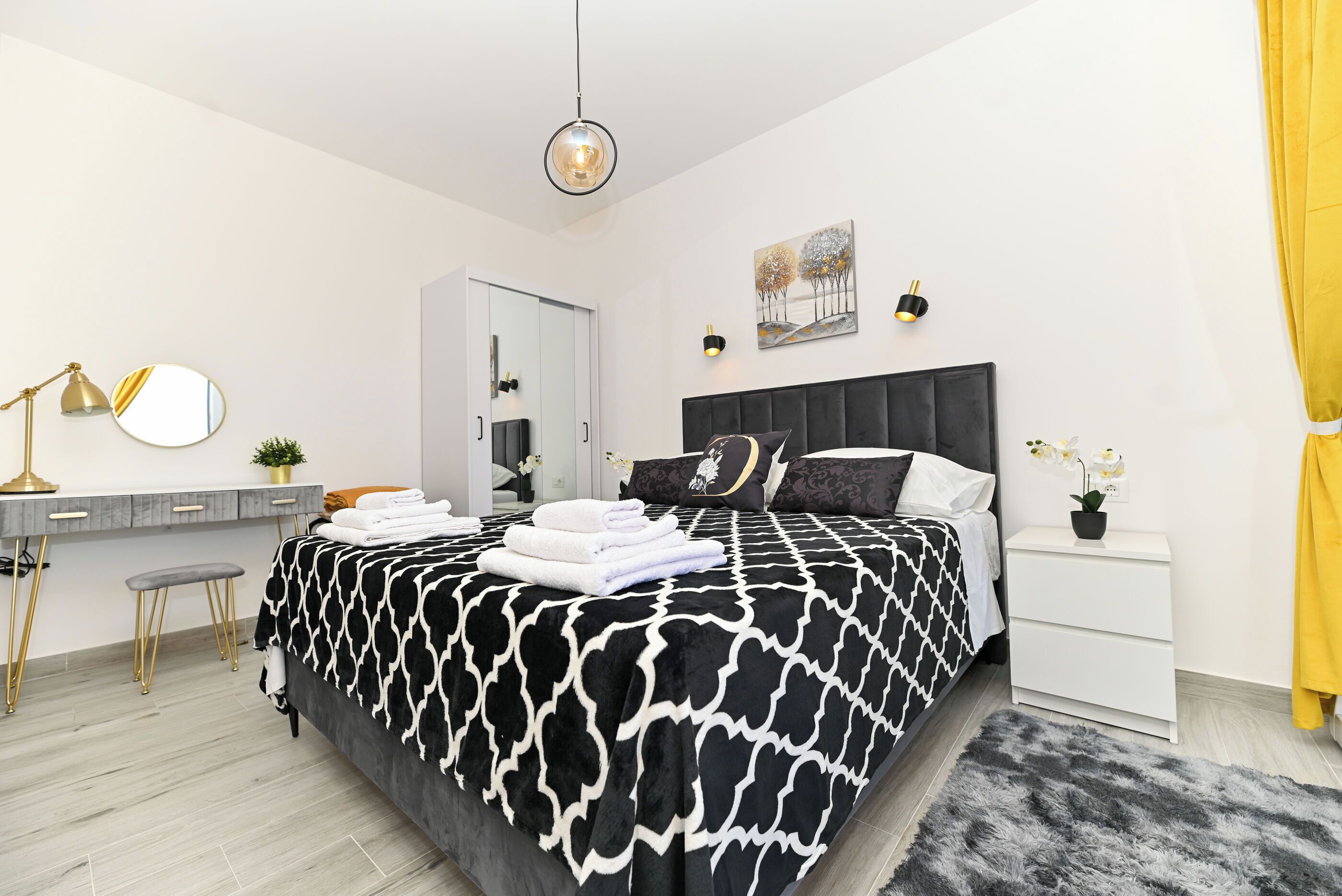 A modern bedroom with a bed, bedside table, nightstand, and a desk with a lamp.