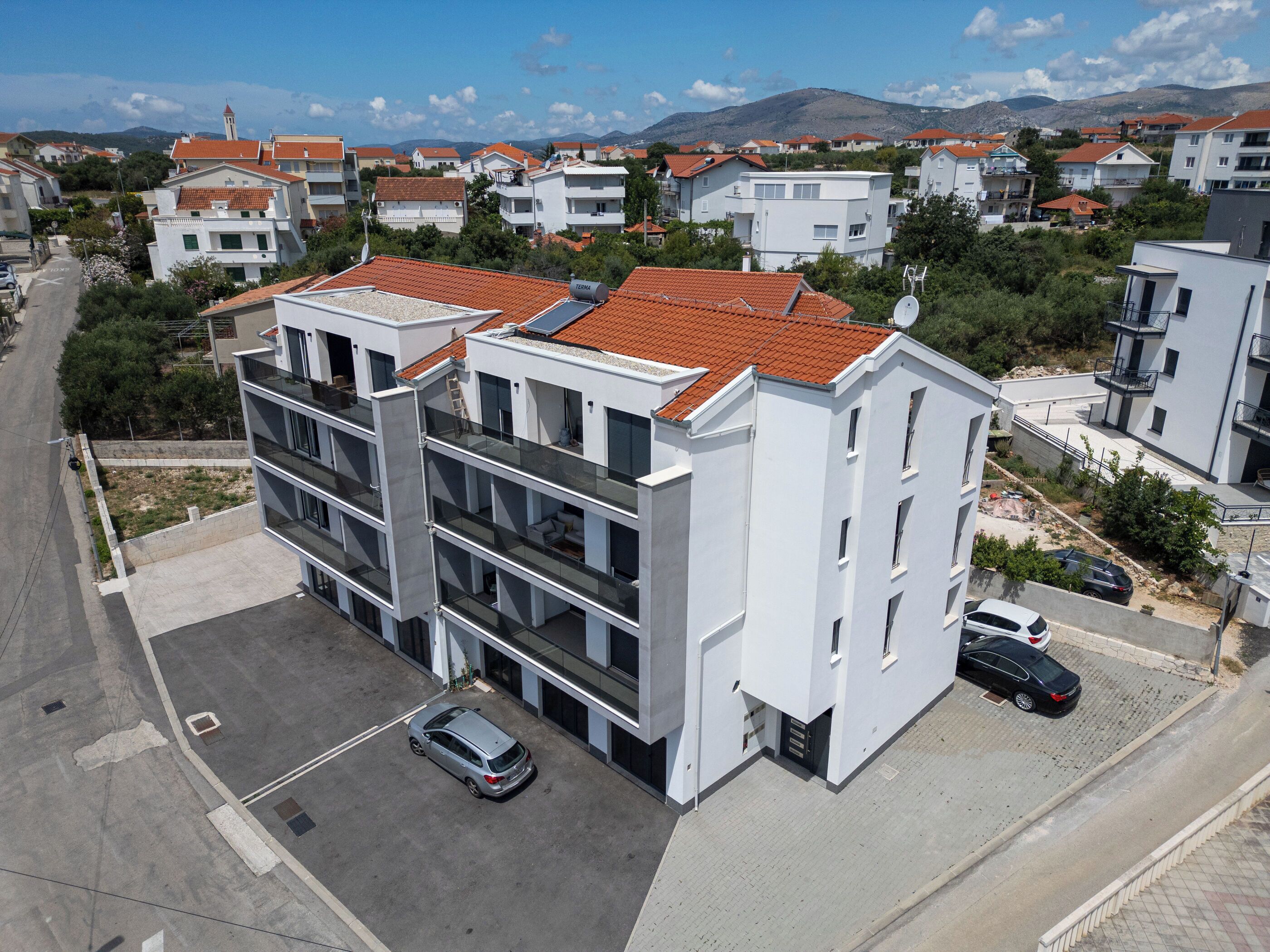 A multi-story residential building with a red-tiled roof, balconies, and a parking area.