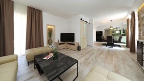 A modern living room with a sofa, a coffee table, and a TV. There is a dining area with chairs and a table. The kitchen is visible in the background with green cabinets. - House Bona (Sukosan)