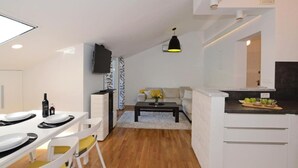 A modern kitchen with white cabinetry, a dining area with a white table and chairs, a sofa, and a black pendant light. - Apartment Leo (Ploce)
