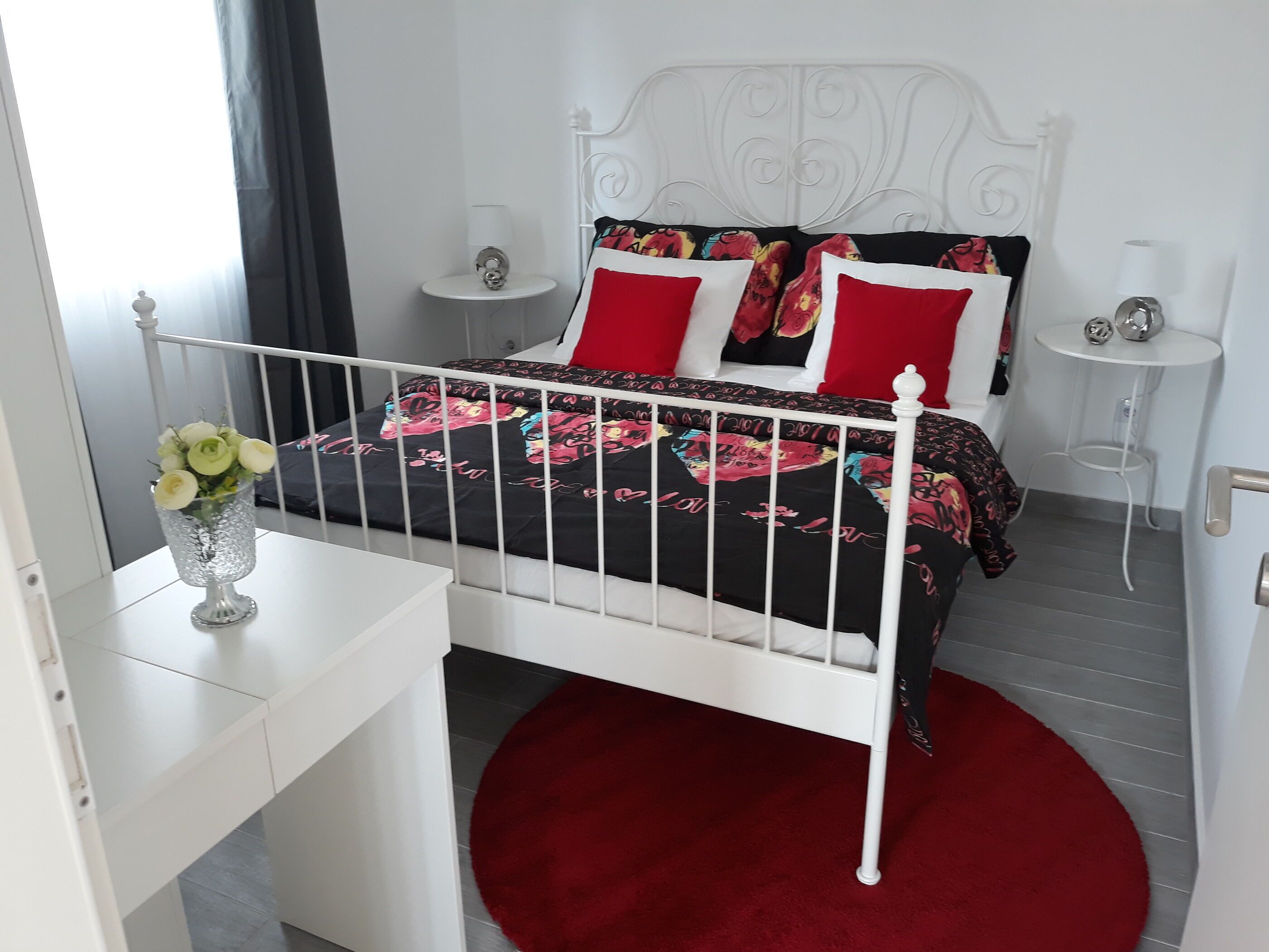 A modern bedroom with a white metal bed frame, a red rug, and a white bedside table with a vase of flowers.