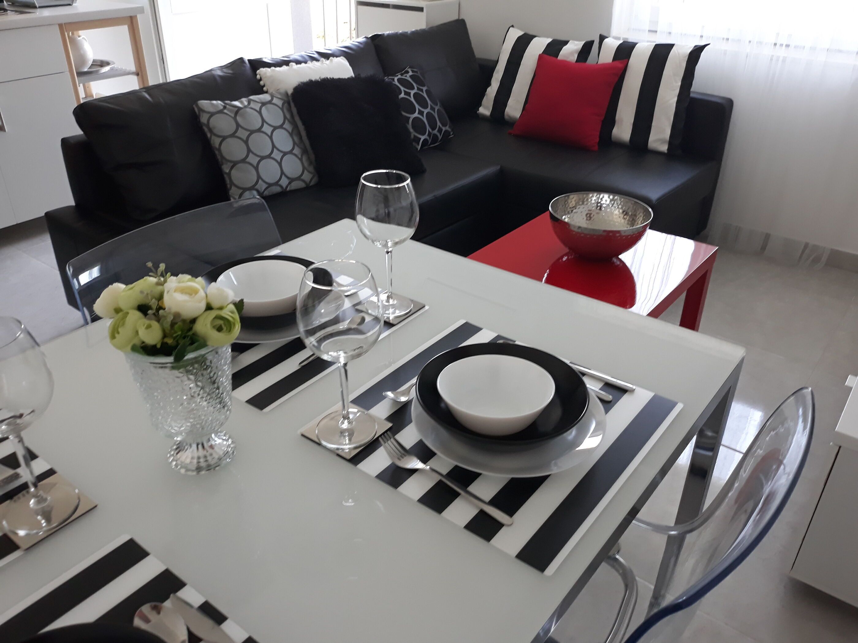 A dining area with a white table set for two, featuring black and white striped placemats, clear glassware, and a vase with white flowers.