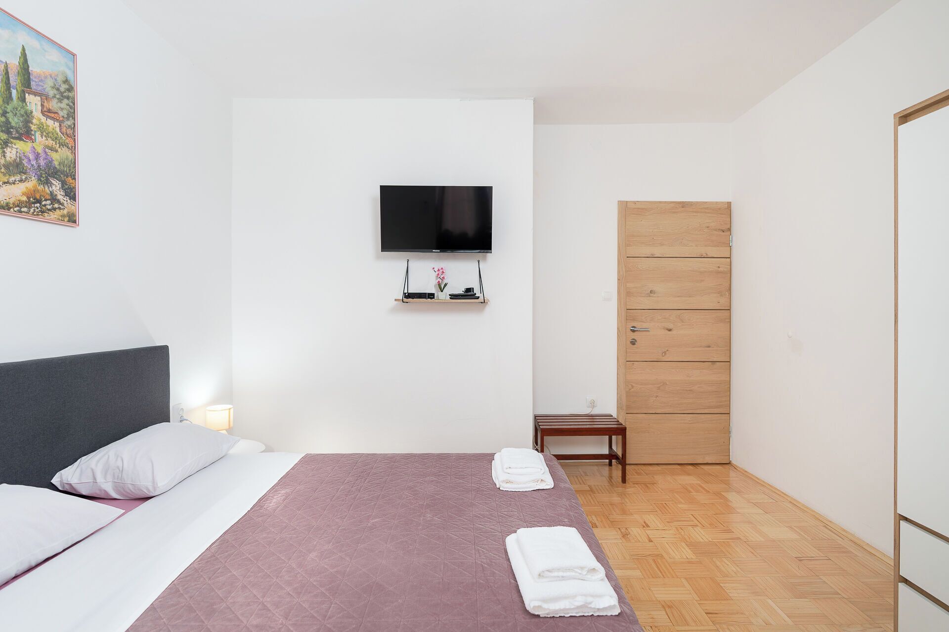 A bedroom with a bed, a TV mounted on the wall, a wooden wardrobe, and a small table.