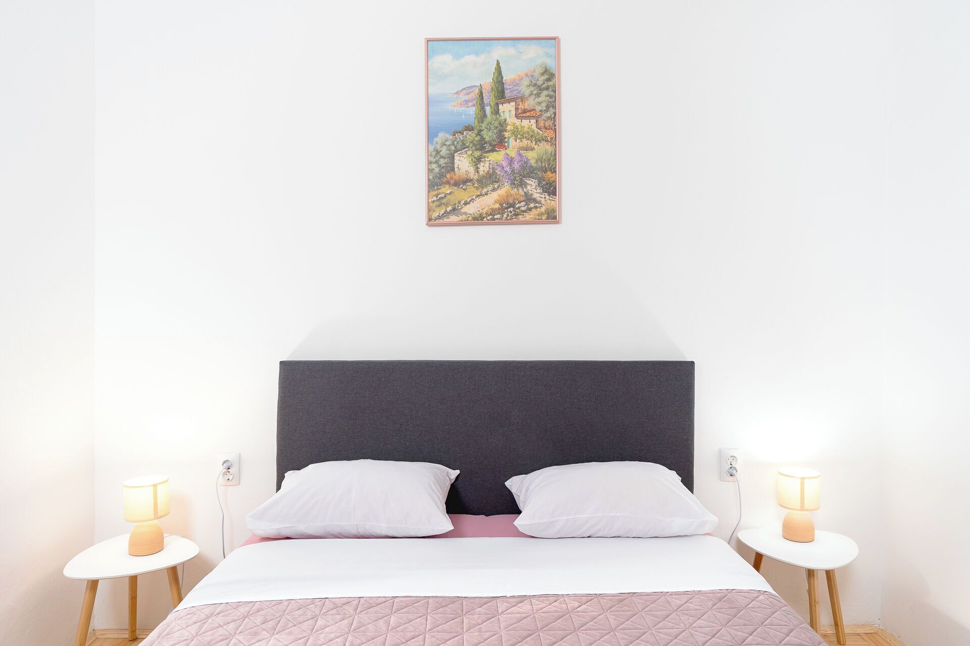 A bedroom with a large bed, two bedside tables with lamps, a dark headboard, and a landscape painting on the wall.