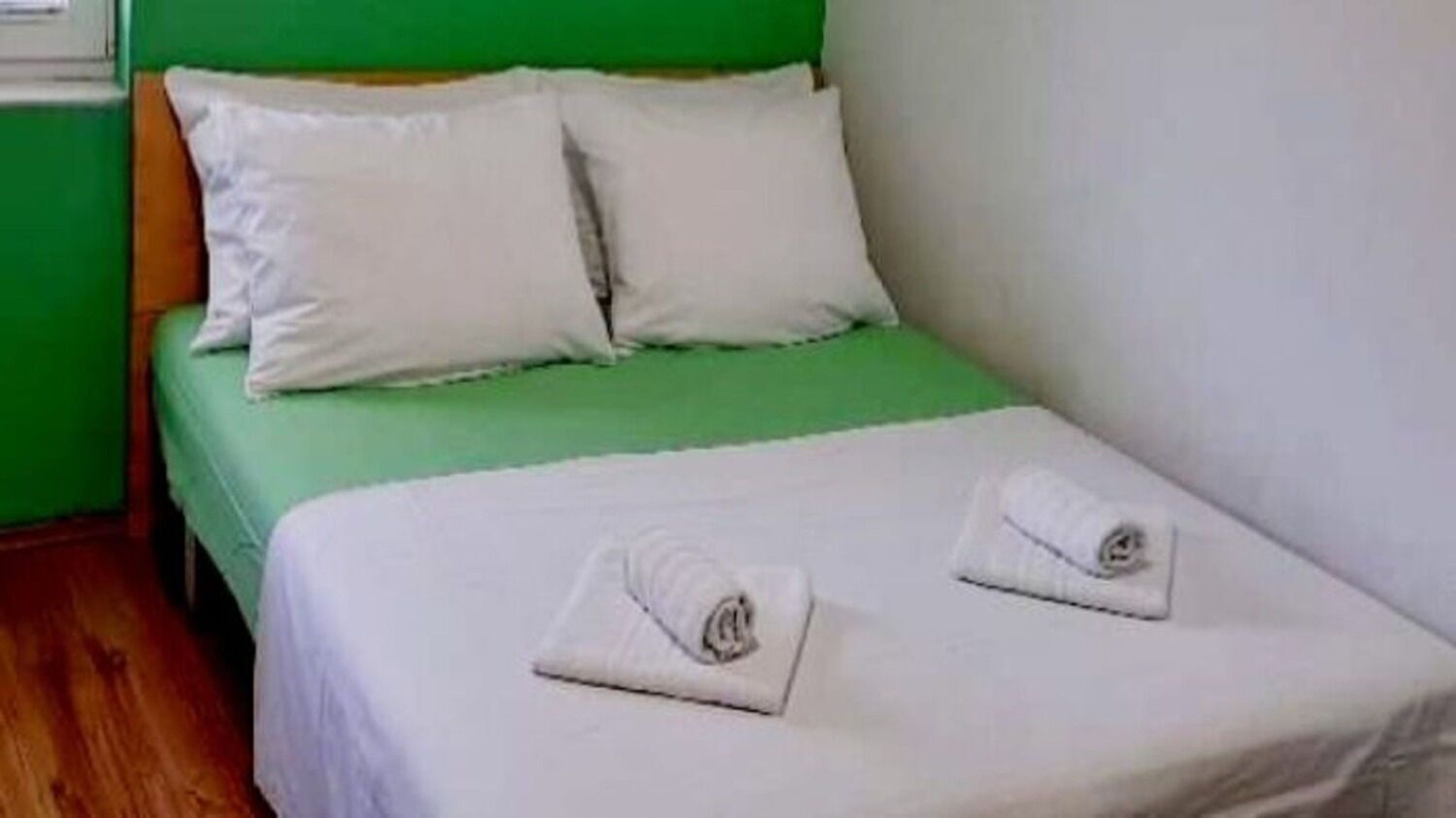 Una cama individual con cabecera verde, ropa de cama blanca y dos toallas enrolladas sobre la cama.
