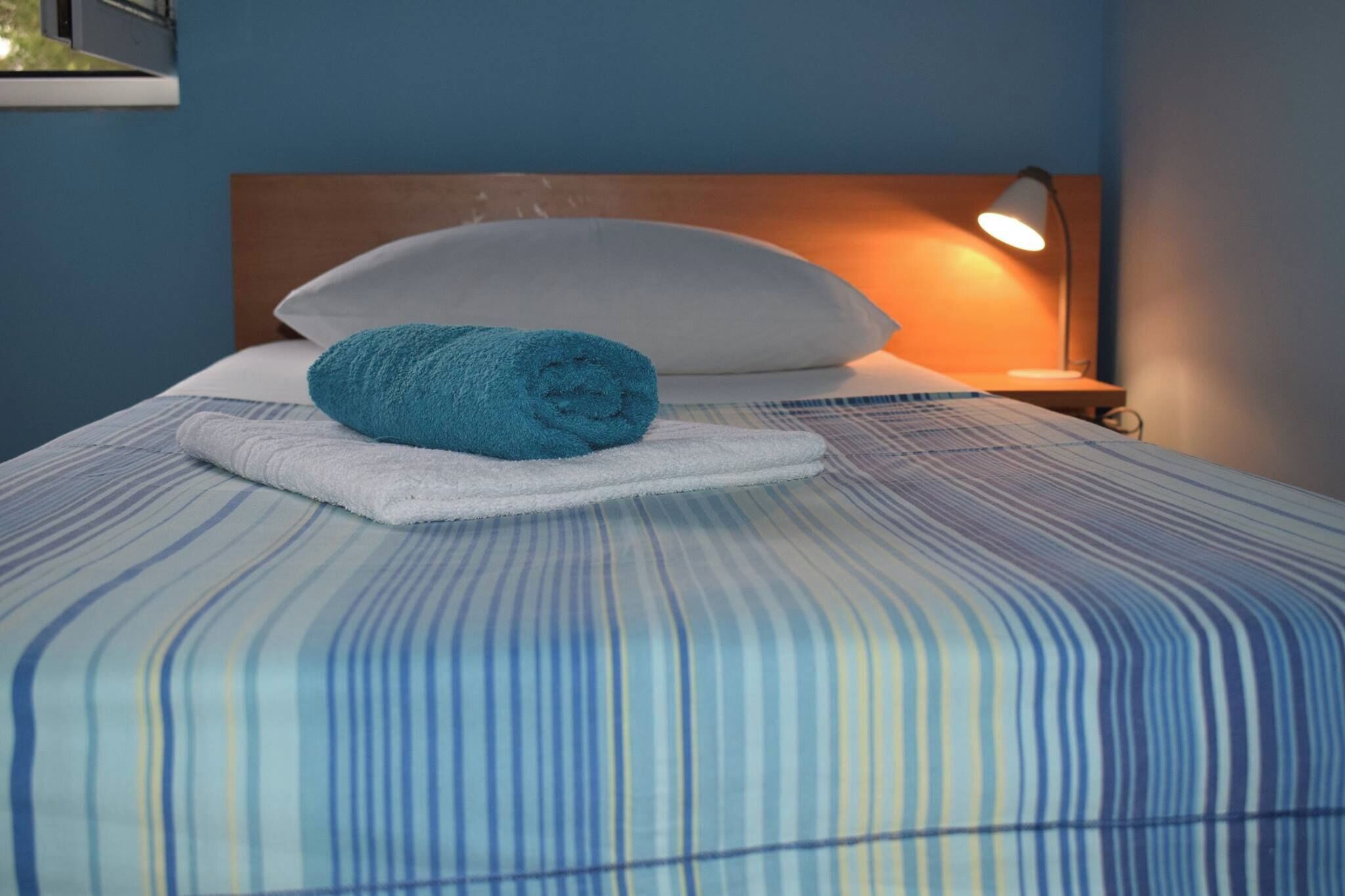 Ein ordentlich bezogenes Bett mit einer gestreiften Tagesdecke, einem zusammengerollten blauen Handtuch und einem weißen Kopfkissen.