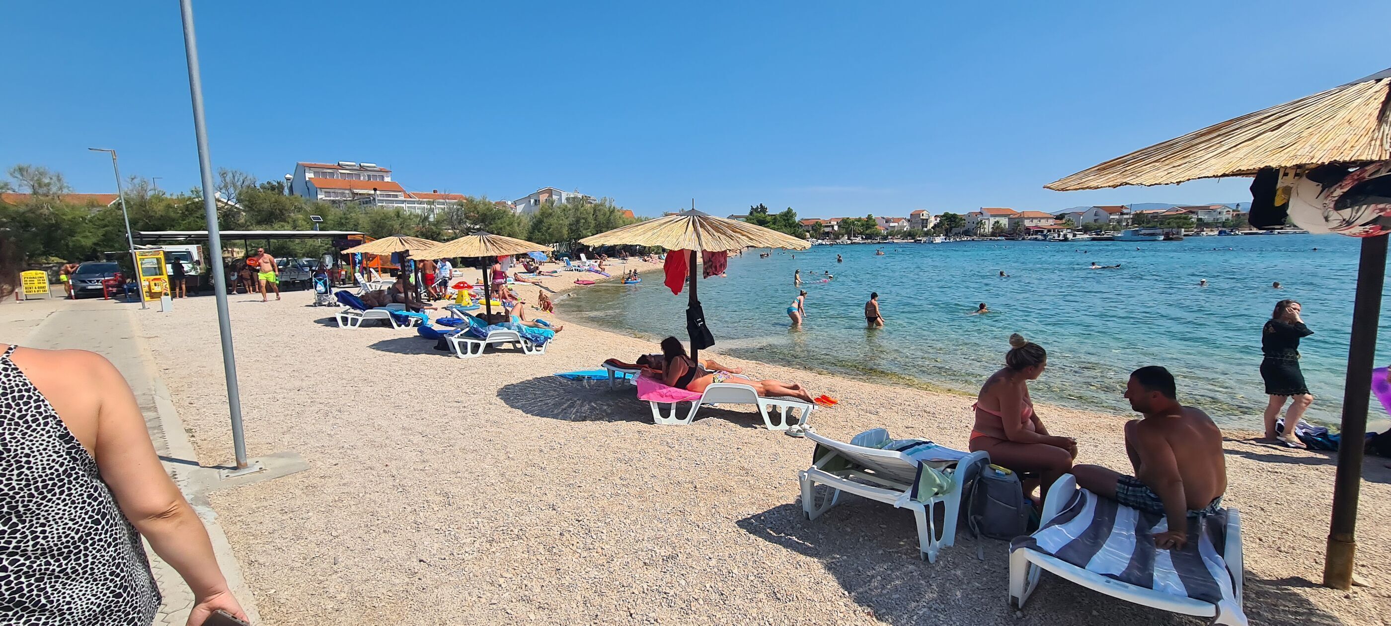 A beach with sun loungers and umbrellas, a clear blue sea, and buildings in the background.