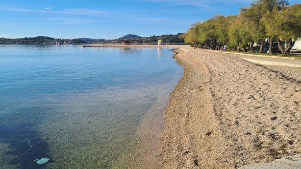 Una playa de guijarros con agua cristalina, un muelle de madera y árboles a lo largo de la orilla.