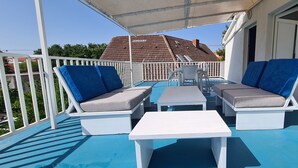 Un espacio de estar al aire libre cubierto con mobiliario blanco y cojines azules, una pequeña mesa y vistas a una casa con techo de tejas.